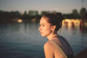 A woman sitting near calm water looking at the sunlight. Women's issues therapy in Lynn, MA can help you explore questions about your identity & disconnection. Get started with a therapist today.