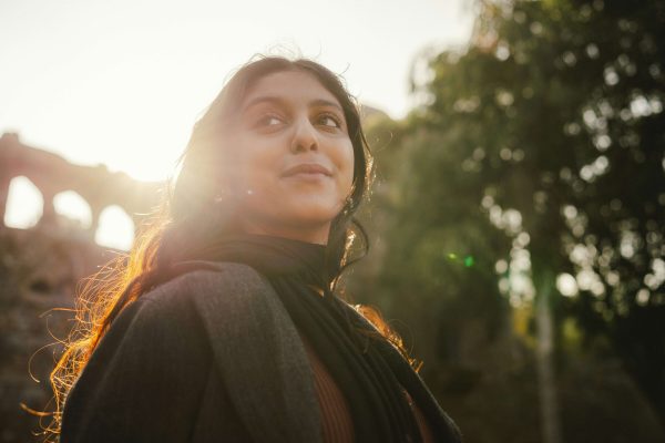 A woman standing outside near trees with the sun reflecting behind her. Women's therapy in Lynn, MA can be done online & in-person for anxiety, identity questions & more. Reach out today for therapy.